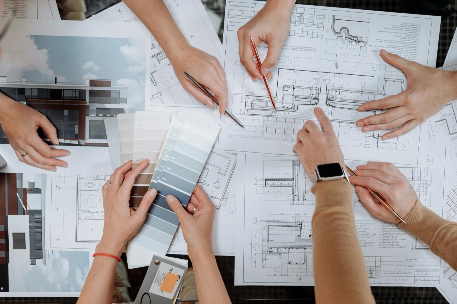 Several pairs of hands working across printed floor plans and elevations spread over a table, one holding a fan of grey and blue material swatches, others holding pencils