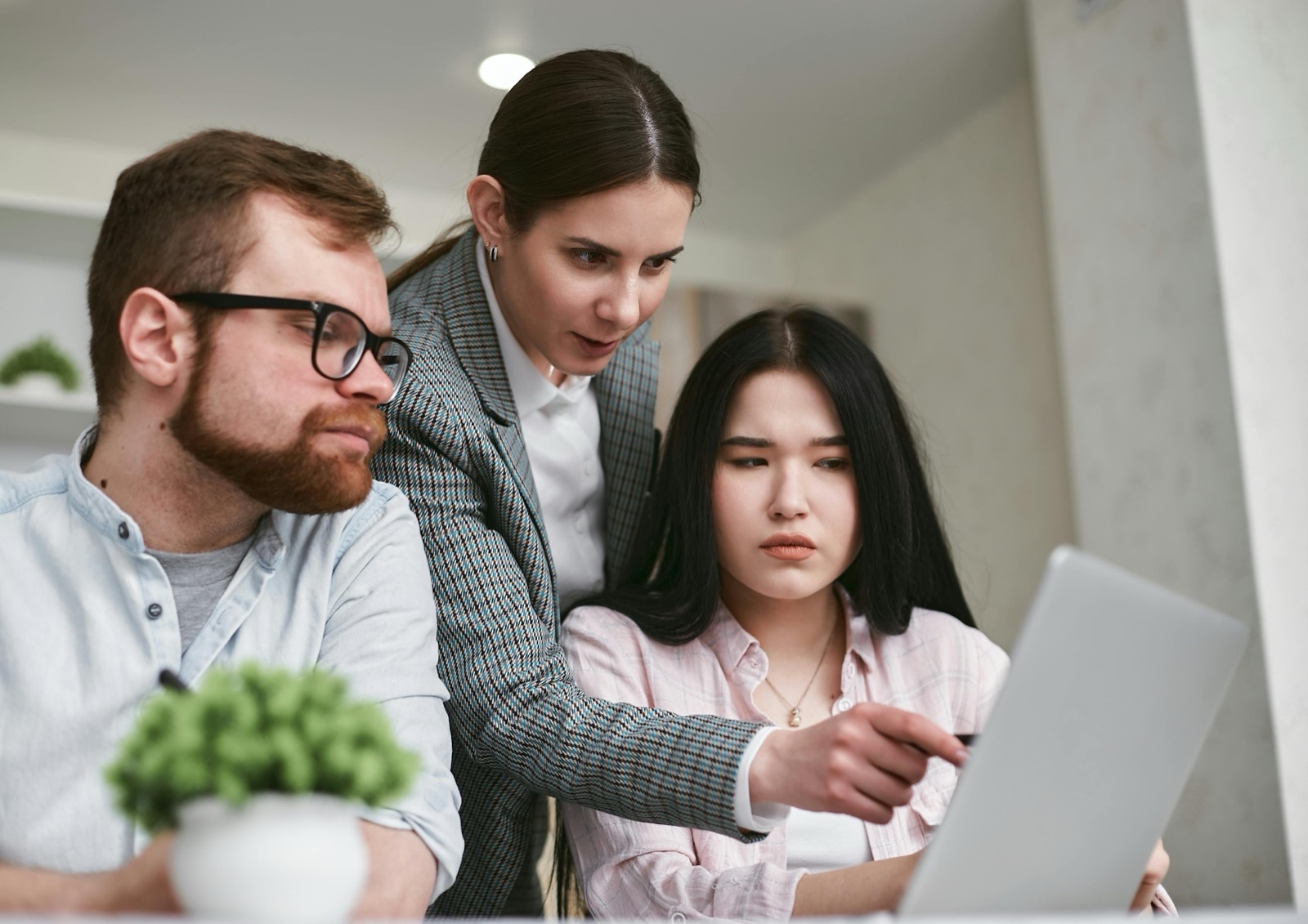 Three colleagues at a desk looking at the same laptop screen, one standing behind the other two and pointing at something on it