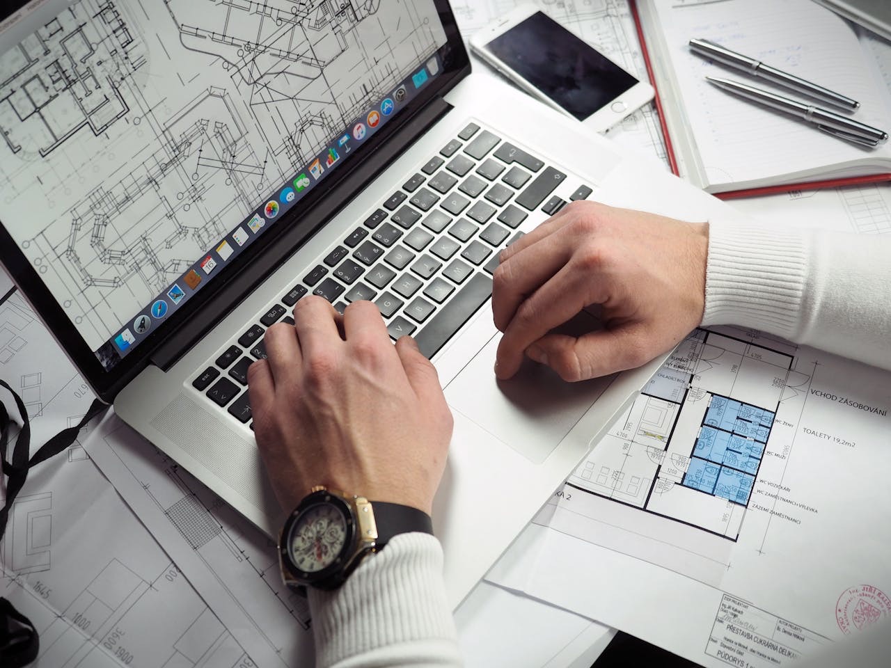 An overhead view of hands working at a laptop showing a CAD floor plan, the desk around it covered with printed architectural drawings, a notebook and pens