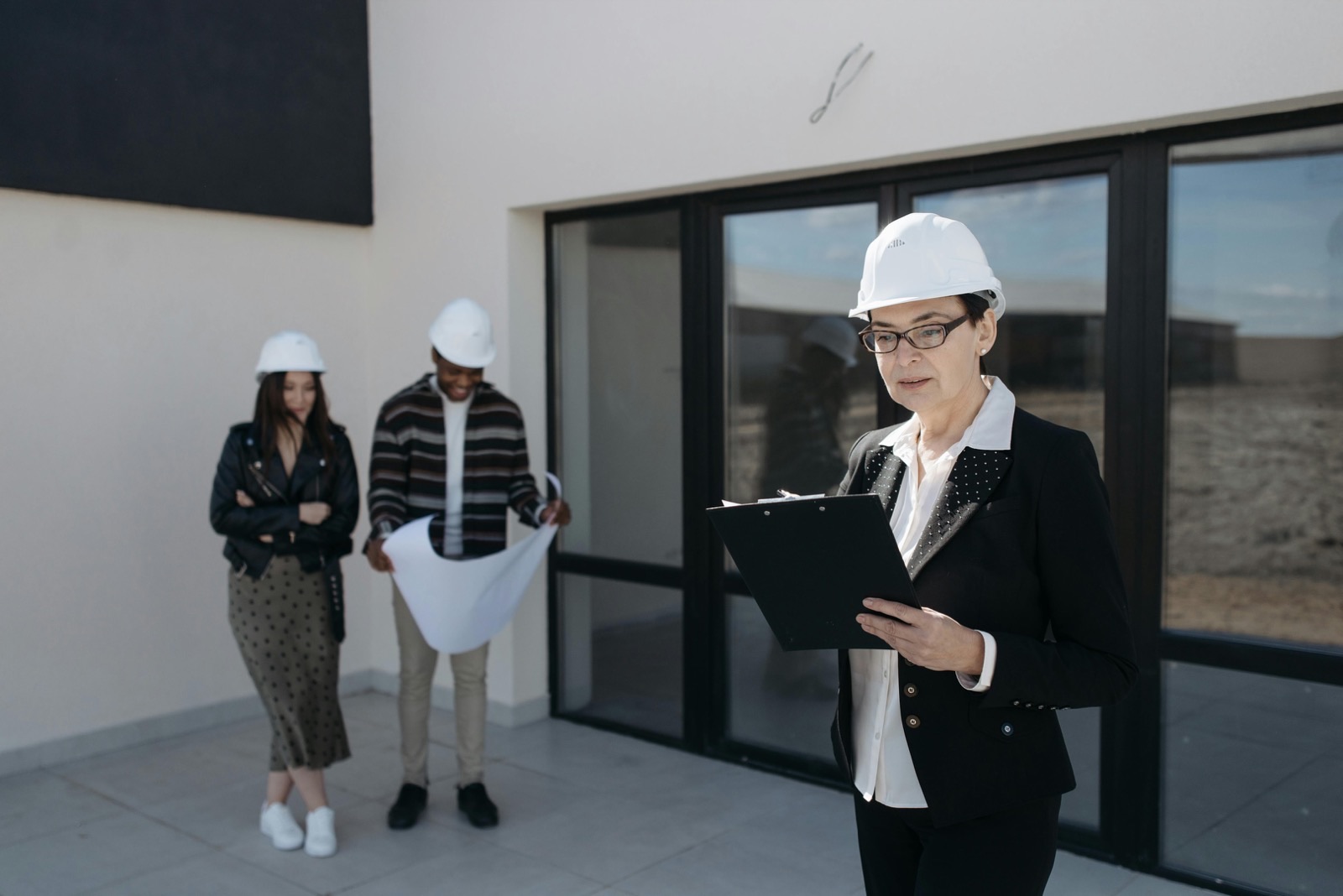 An engineer in a hard hat working down a checklist on a clipboard outside a newly completed building, while two colleagues behind her unroll a printed drawing to compare against it