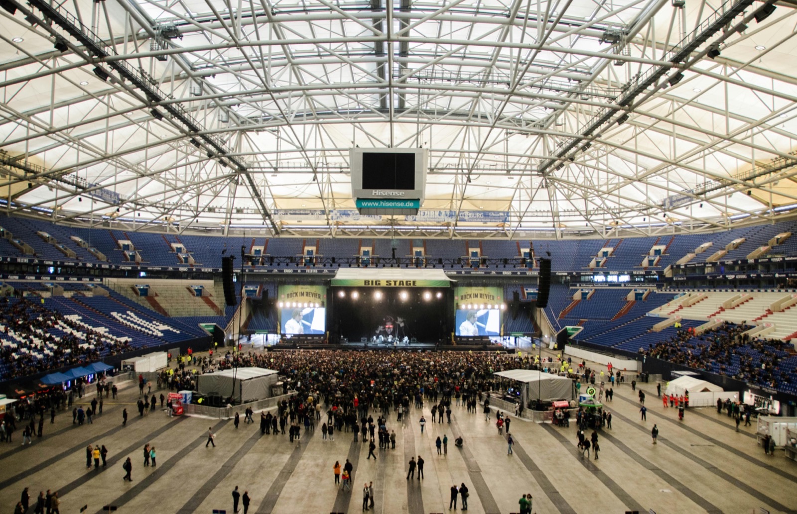 The interior of a covered arena set up for a concert, with a stage at one end, a standing crowd across the floor and a steel roof truss grid overhead