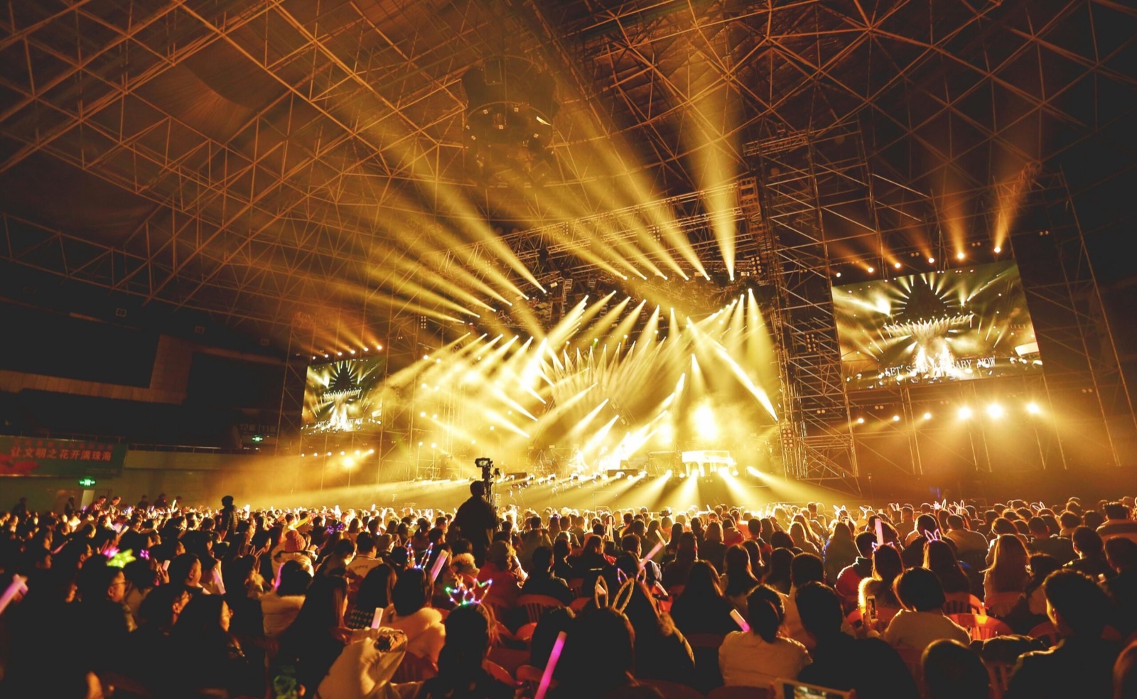 A packed arena during a concert, the crowd on its feet across the whole floor while beams of stage light fan out beneath an exposed steel space-frame roof