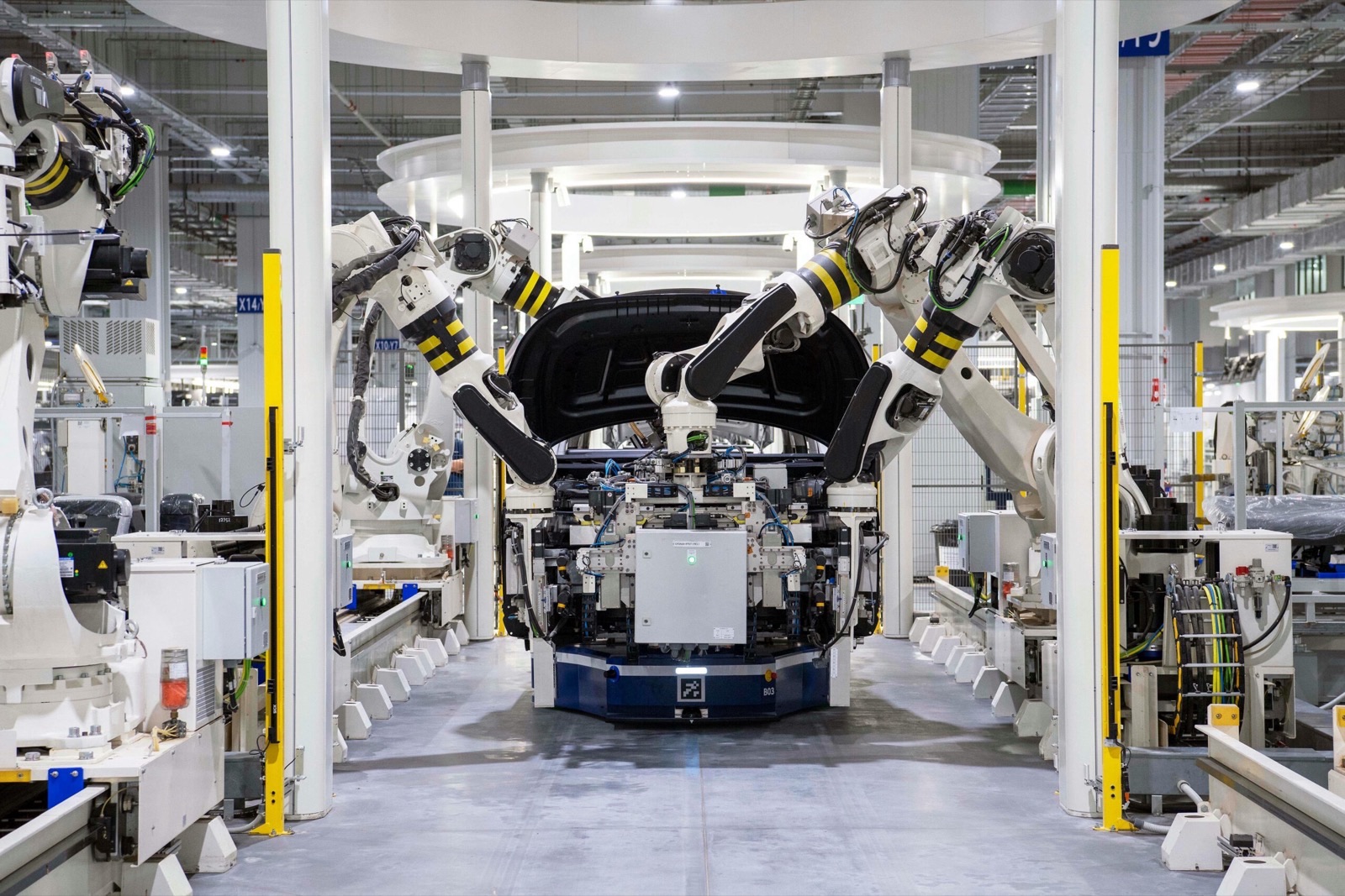 Robotic arms on either side of an assembly line lifting a car body panel over the workpiece below, inside a manufacturing hall with an overhead conveyor ring and yellow guard posts lining the aisle
