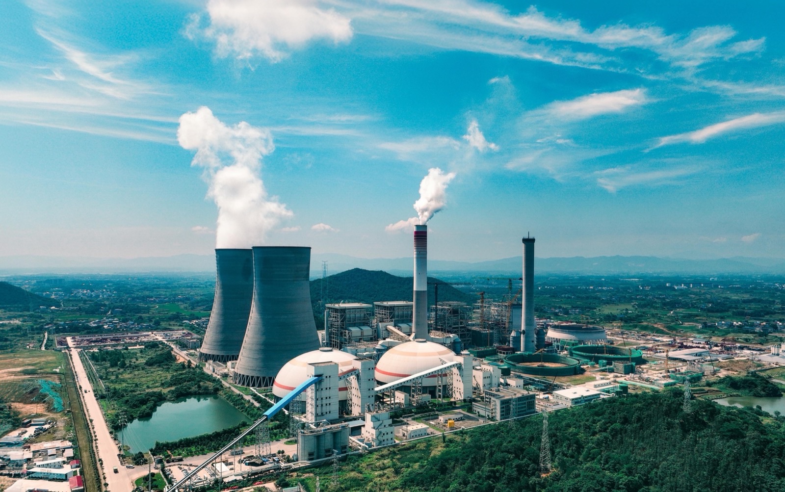 Aerial view of a thermal power station in green hill country: two hyperboloid cooling towers venting steam, a red and white banded chimney stack, domed fuel stores and a switchyard laid out beside them