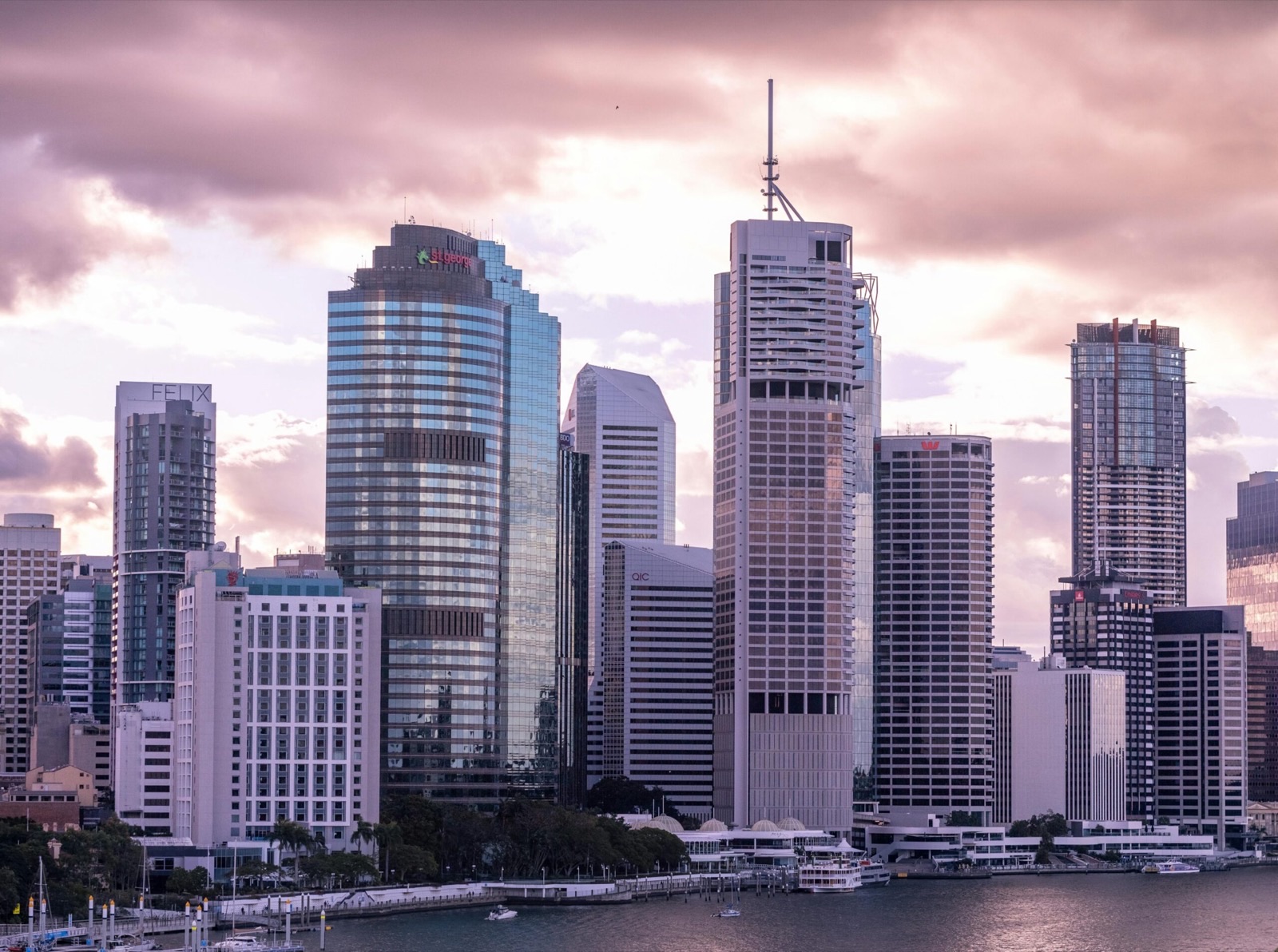 A riverside downtown skyline of glass and concrete office towers under a pink evening sky, one of them still topped out with construction hoarding
