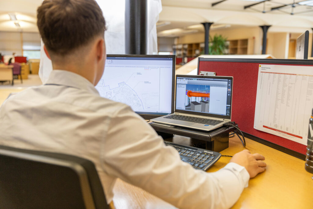 An engineer at a desk with a site layout drawing open on one screen and a 3D coordination model on a laptop beside it, a printed element schedule pinned to the partition