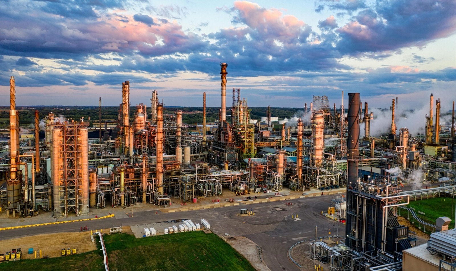 An aerial view of a petrochemical plant at dusk: distillation columns, dense pipework, storage vessels and flare stacks laid out across the site