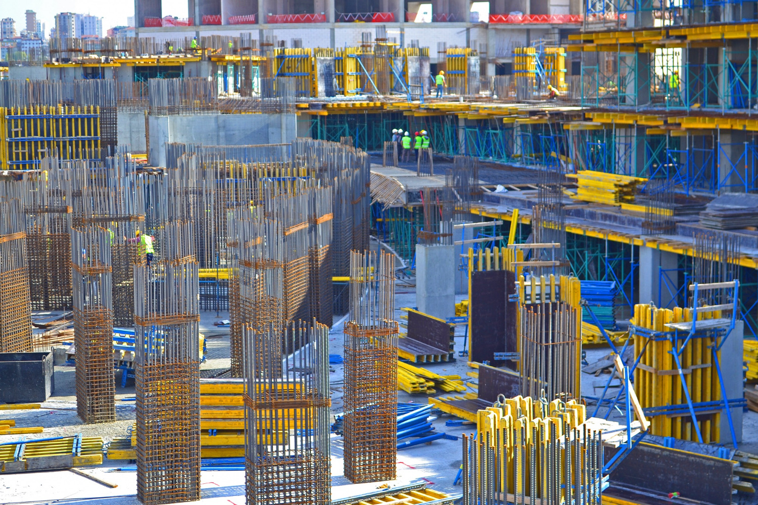 Rows of vertical column reinforcement cages standing in formwork across a multi-level construction site, with scaffolding and stacked shuttering panels behind them