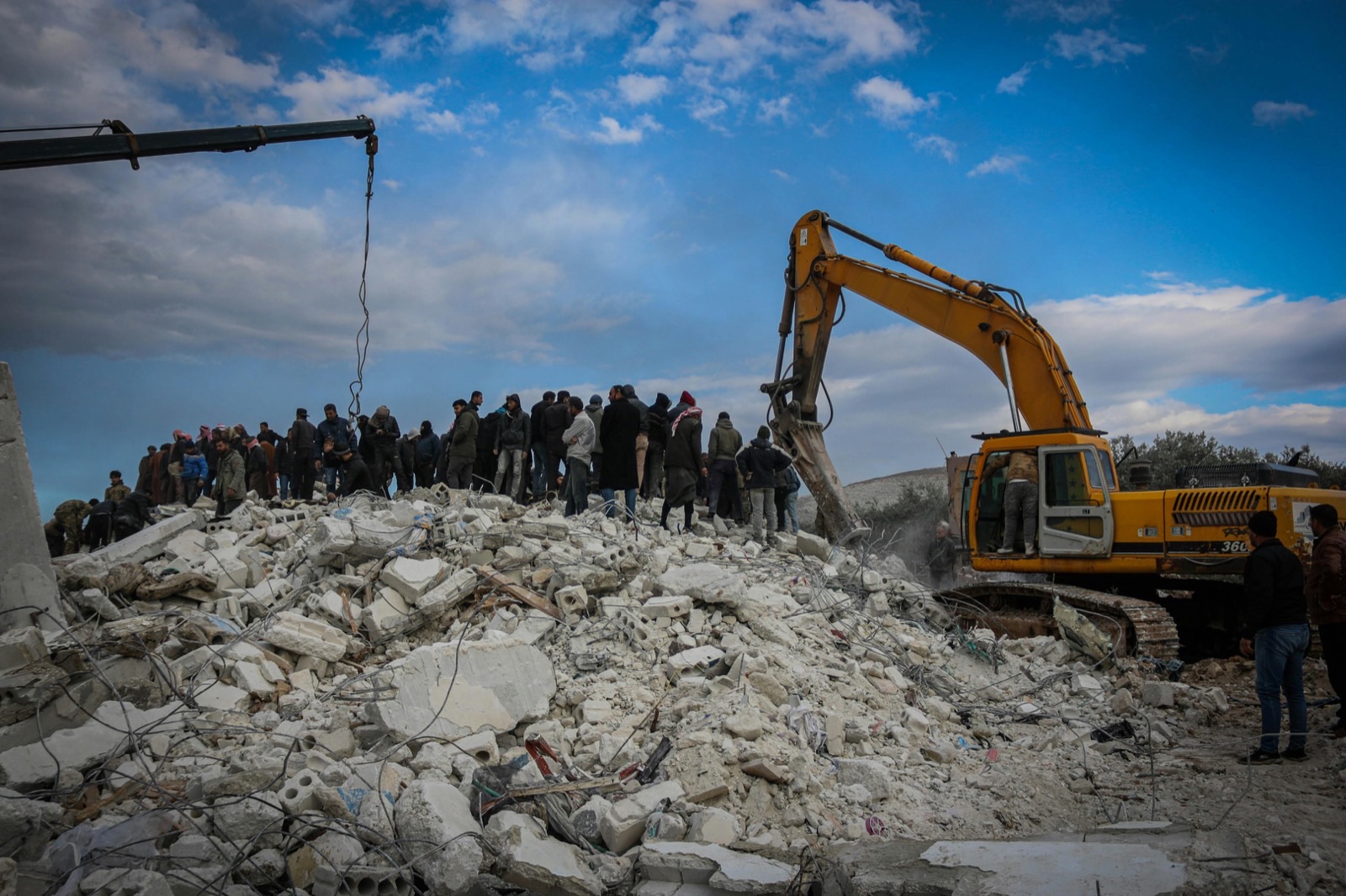 People standing on the rubble of a collapsed concrete building while an excavator works at the debris beside them