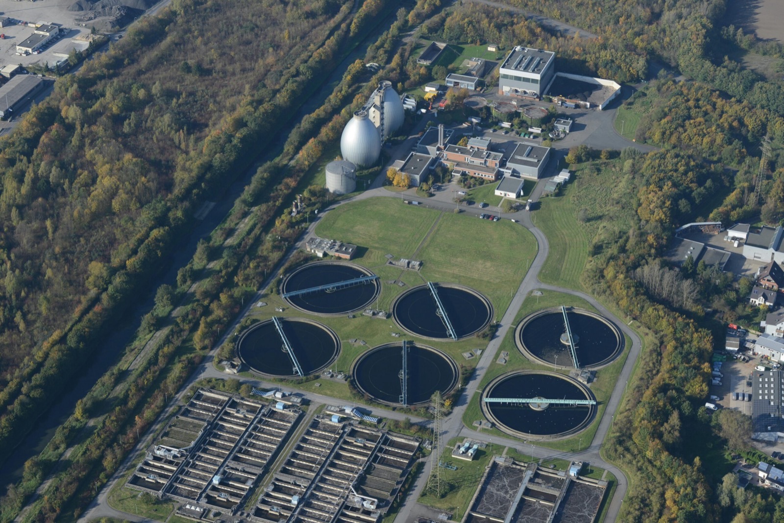 A wastewater treatment works seen at an angle from the air: six circular clarifiers with rotating scraper bridges, banks of rectangular aeration lanes below them, and an egg-shaped digester and silos beside the plant buildings