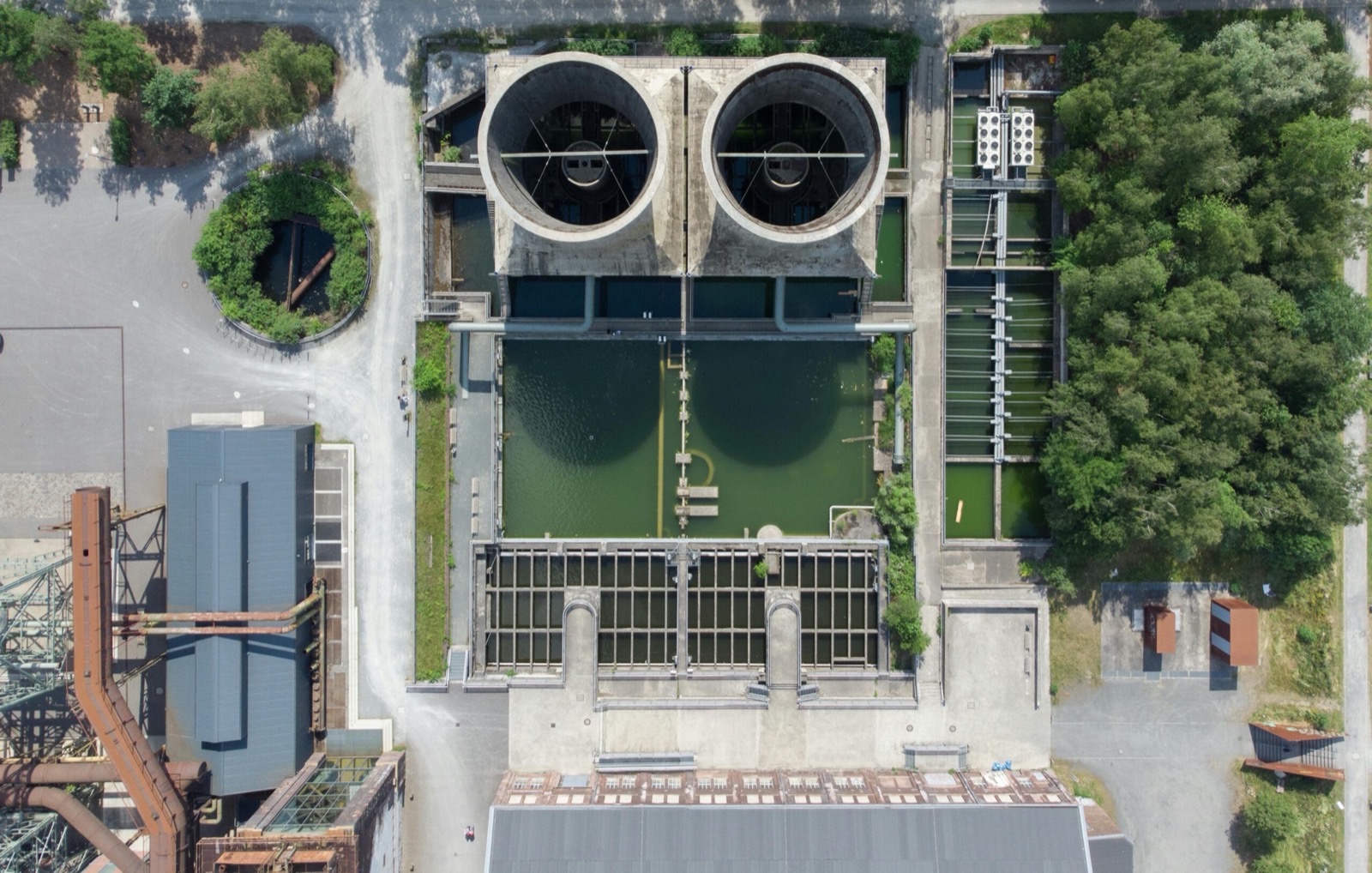 A water treatment structure photographed straight down: two large circular concrete chambers side by side, an open rectangular basin of green water below them crossed by walkways, and weirs and channels feeding between the two
