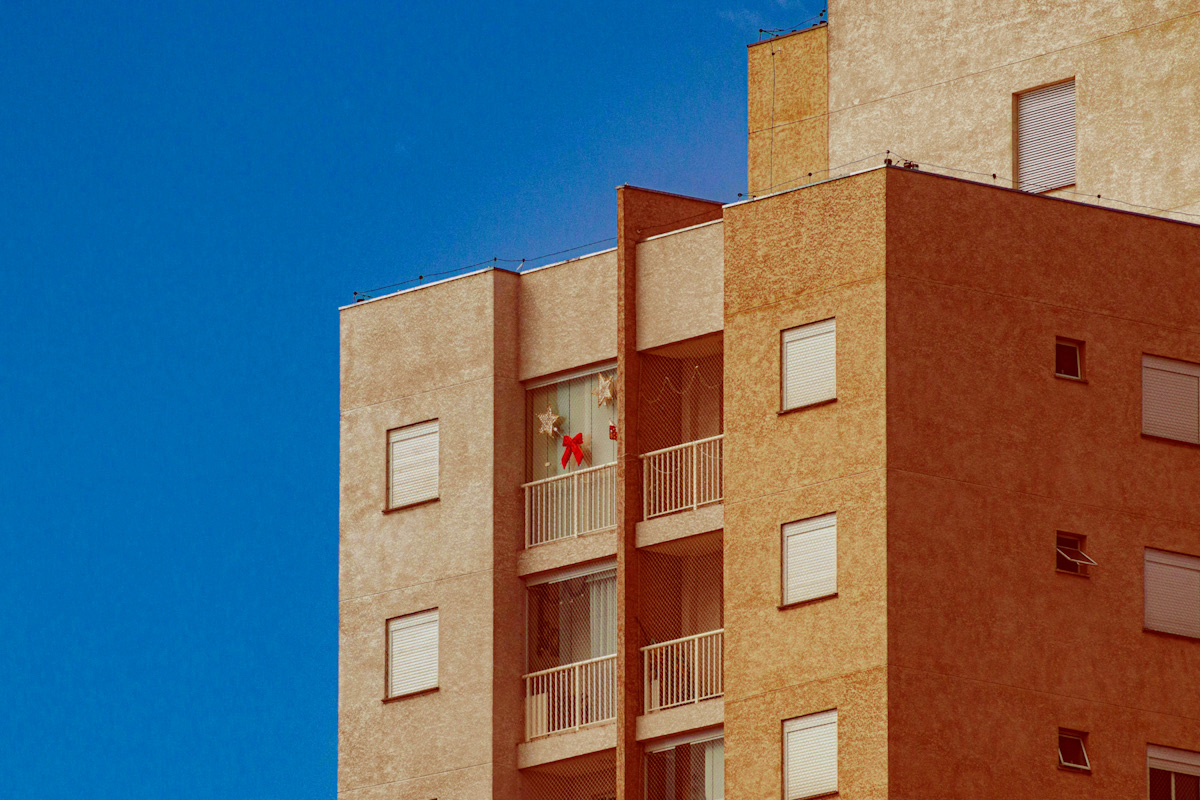 The upper corner of a multi-storey apartment block, its shuttered windows and a recessed balcony bay catching low sun against a clear blue sky