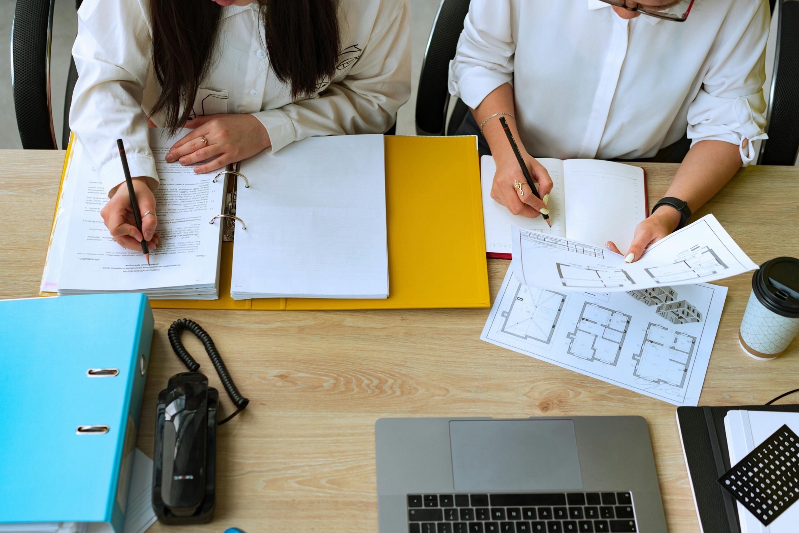 Two people at a shared desk seen from directly above, one annotating a ring-bound document and the other holding a printed sheet of floor plans over an open notebook