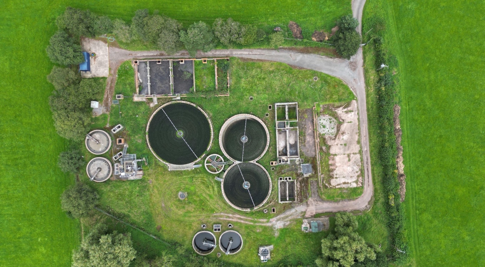 A small rural sewage treatment works photographed straight down from the air: three circular clarifiers with rotating bridges, rectangular tanks and a gravel access track, the whole plant surrounded by green fields