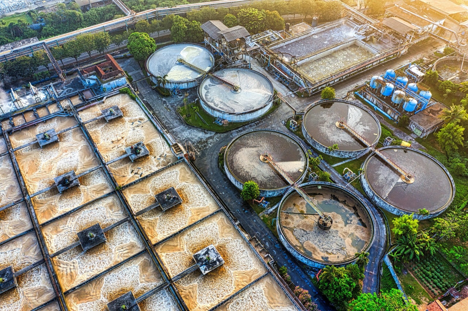 An industrial treatment plant photographed from above at sunrise, foaming rectangular aeration basins filling the left of the frame and circular clarifiers with their scraper bridges on the right, ranks of blue tanks behind them