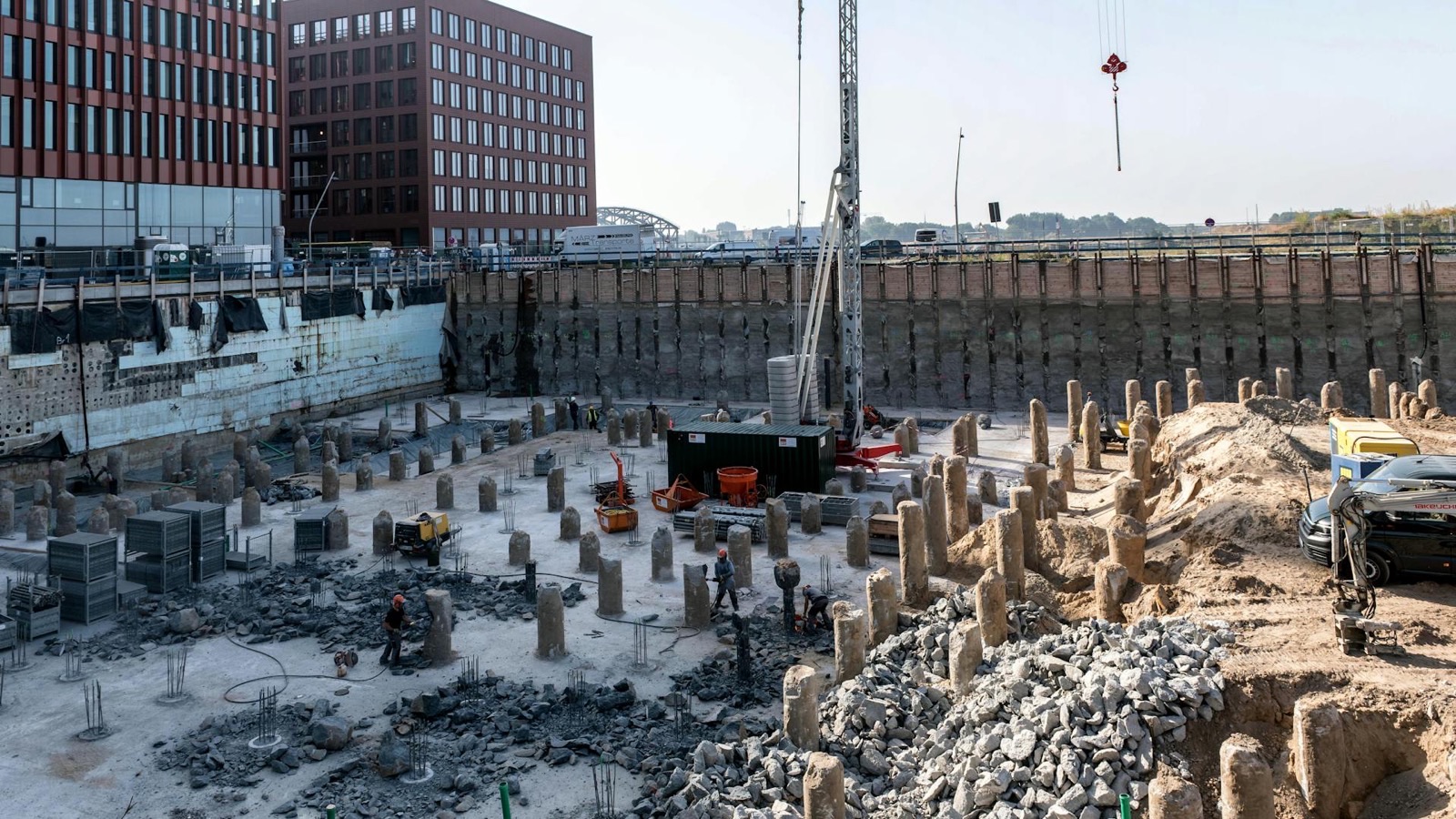 A deep excavation held back by a sheet piled wall, its floor covered with trimmed concrete pile stumps and exposed reinforcement, a piling rig working at the centre