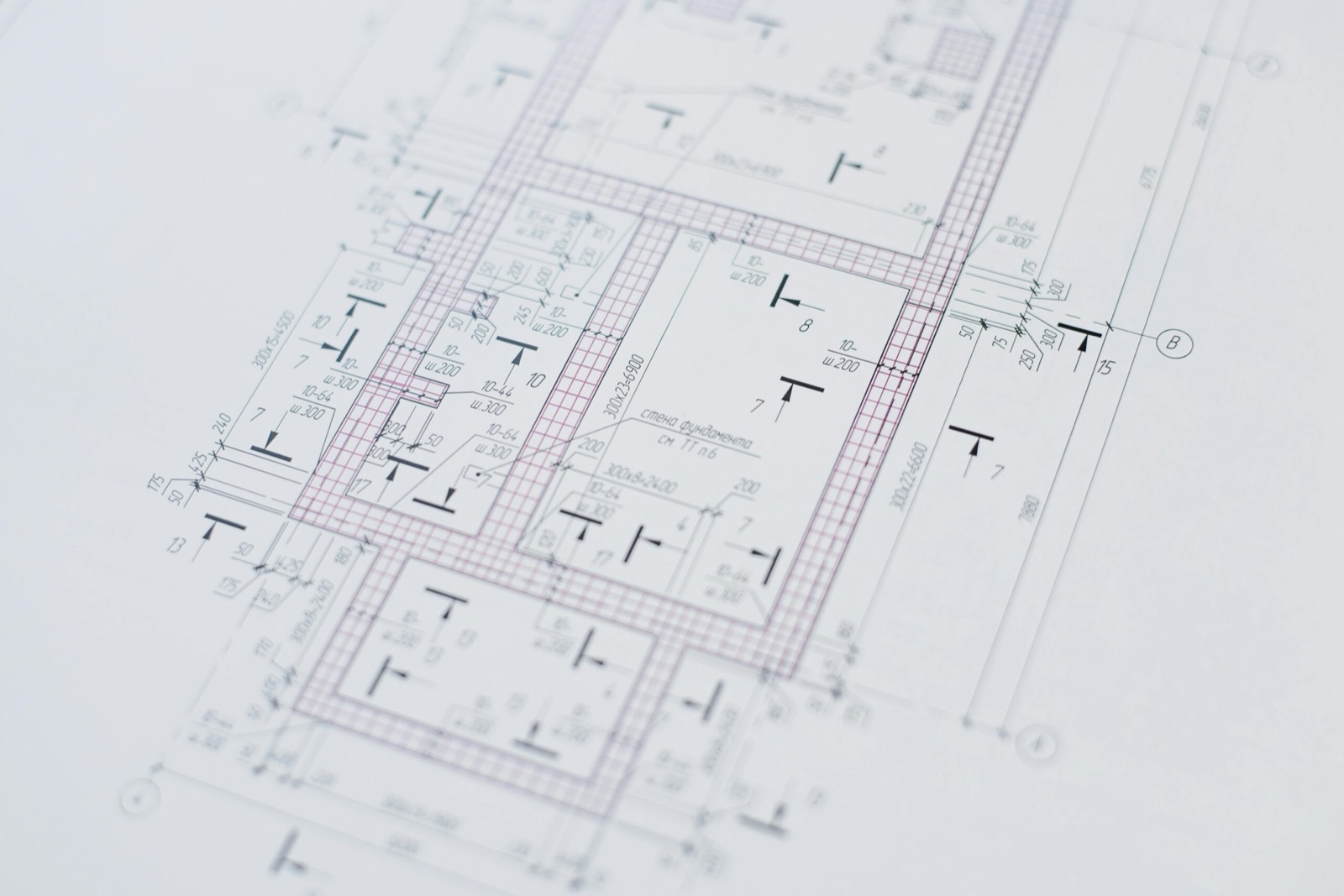 A printed structural foundation drawing lying flat, seen close up at a shallow angle: reinforcement hatching over the footings, dimension strings along every face, section marks and lettered grid bubbles running off the edge of the sheet