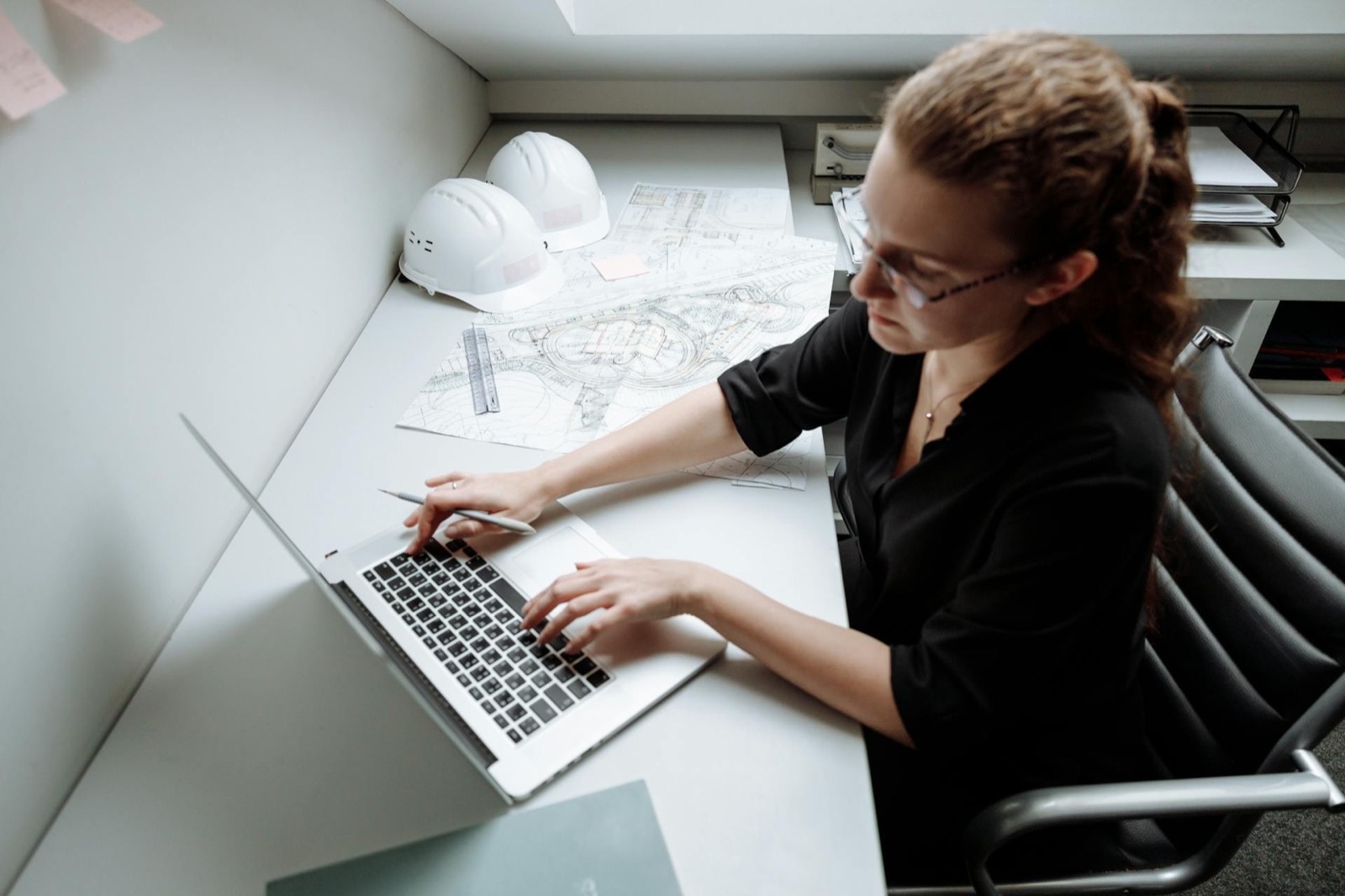 An engineer typing at a laptop on a desk spread with printed site drawings, two hard hats set aside at the end of the bench