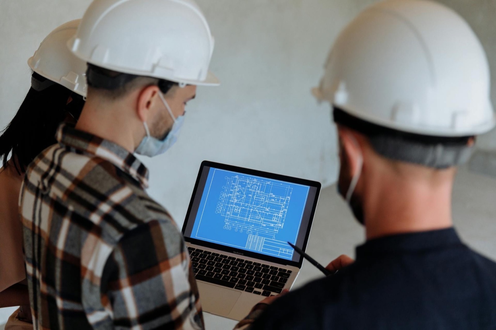 Two workers in hard hats on a bare concrete floor study a laptop showing a blueprint, one pointing at the screen with a pen