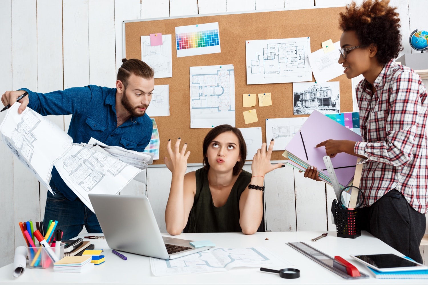 Two colleagues lean over a desk to challenge a third, loose printed floor plans flying above a laptop, a pinboard of drawings behind them