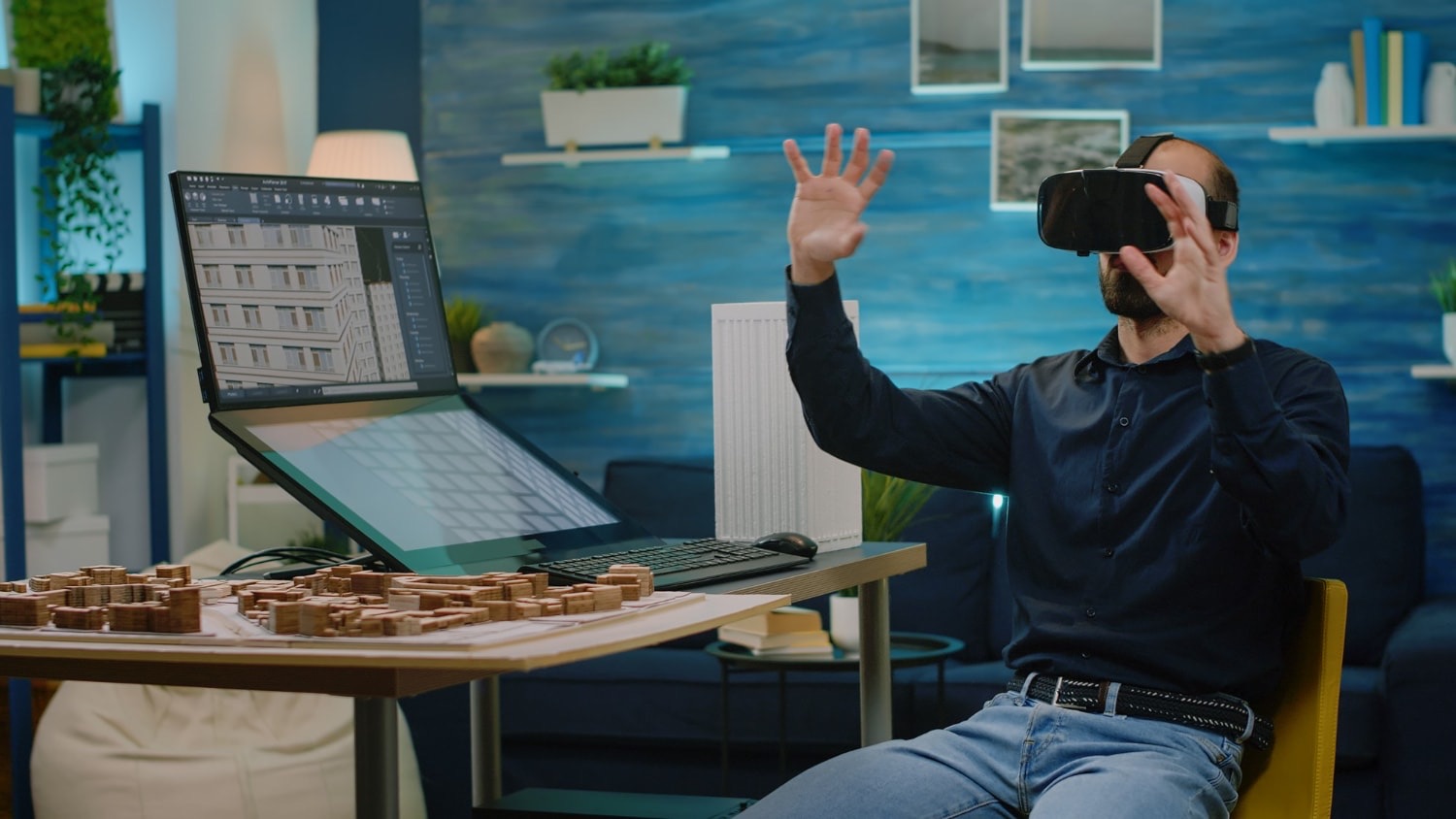 A designer wearing a virtual reality headset and reaching into the air at his desk, a building model open on the monitor beside him and a physical scale model of the same city block laid out on the desktop