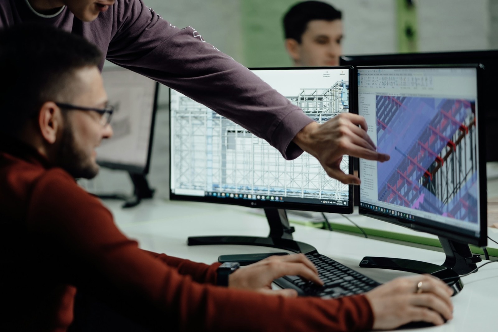 Two engineers at a desk of paired monitors, one leaning in to point at a 3D structural frame on the left screen while the right screen shows the same model colour-coded beside a properties list