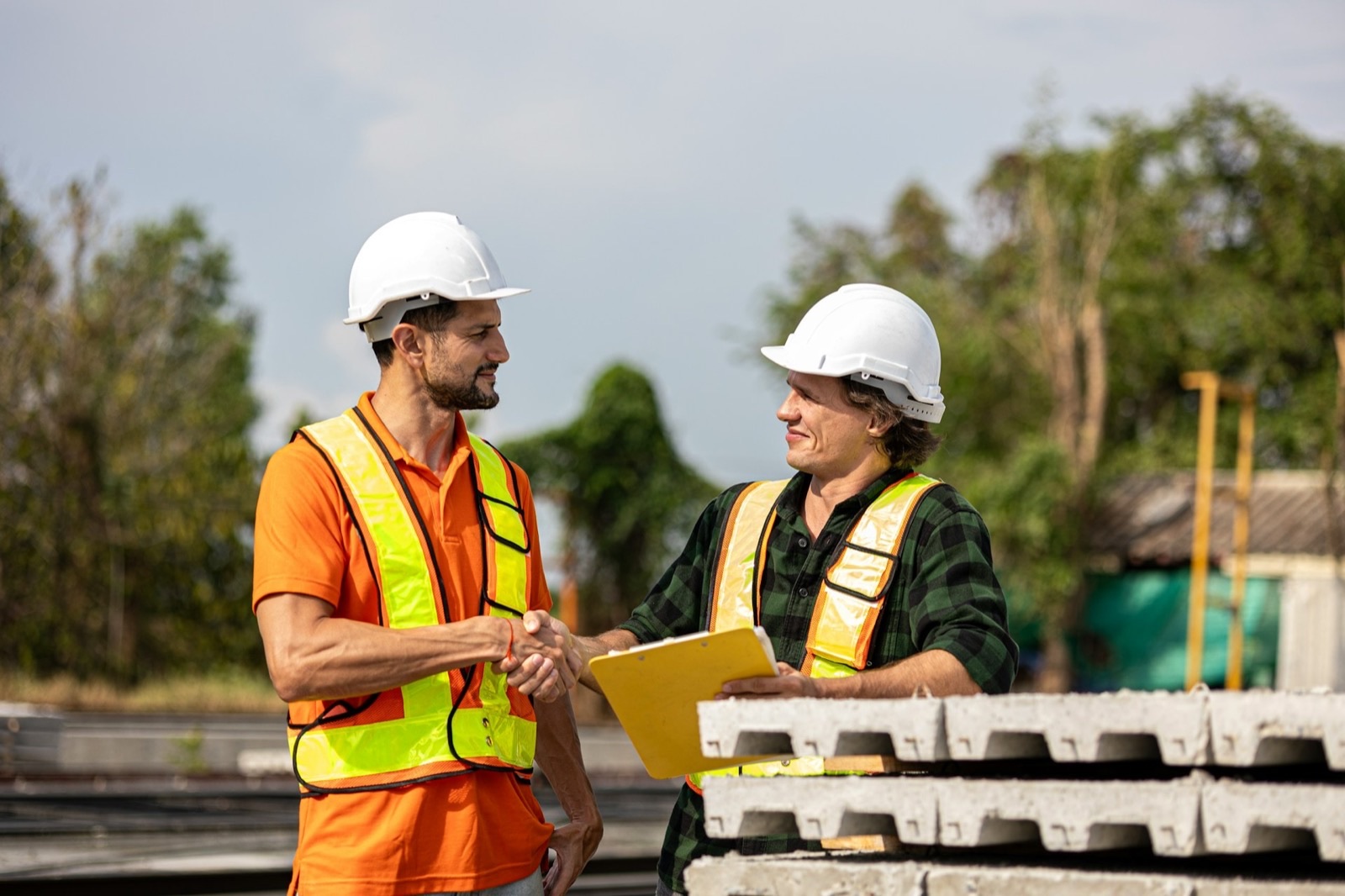 Two workers in hard hats and high-visibility vests shaking hands beside a stacked pile of ribbed precast concrete units, one of them holding a yellow clipboard