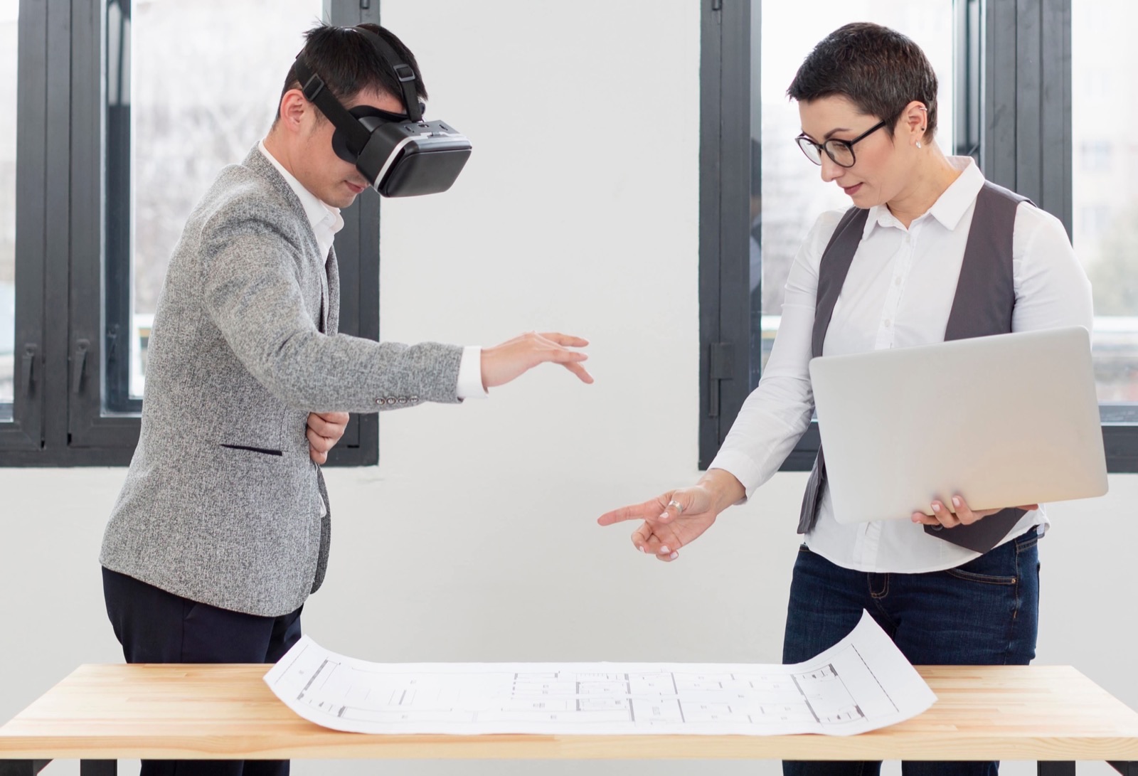 A person wearing a virtual reality headset reaching into the air while a colleague holding a laptop gestures toward a large drawing unrolled on the table between them